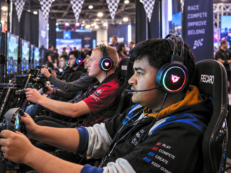 Students racing side by side in sim rigs at an esports expo, headsets on, hands on the wheel.