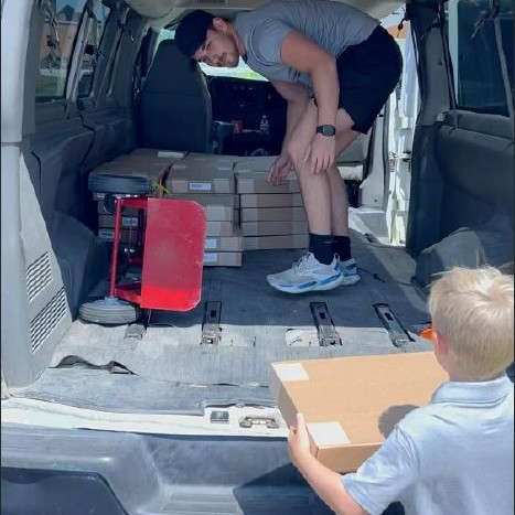 Boxes of refurbished computers being unloaded from a van, with a young student watching.