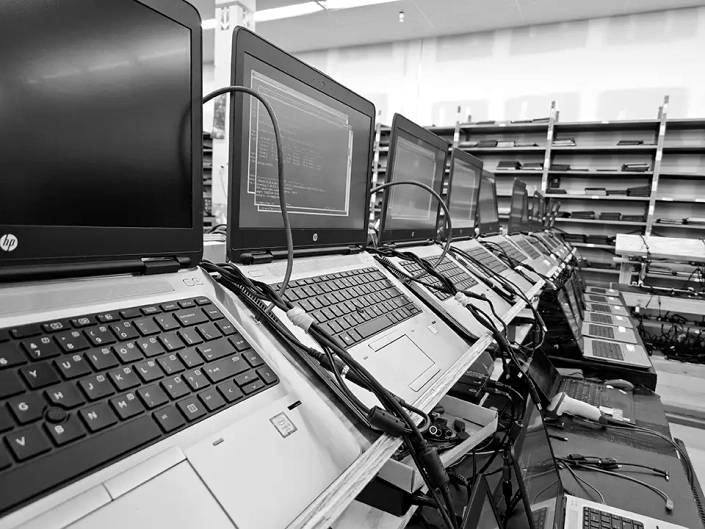 A long bench of open business laptops on the Regadget floor, each one cabled up for testing.