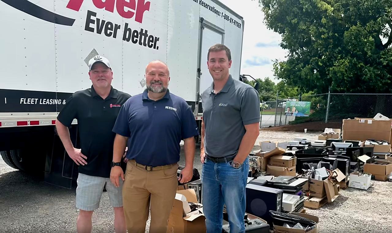 Three people standing by a box truck at a tech drive, with collected computers, monitors and boxes stacked on the ground beside them.