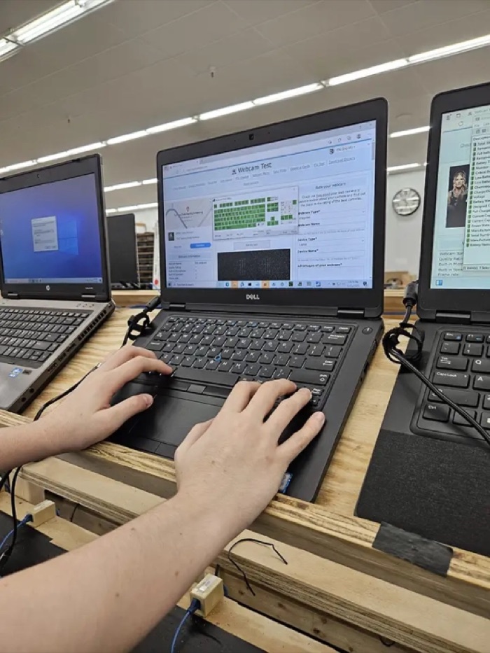 A technician runs a keyboard and webcam test on a refurbished Dell laptop at a testing bench.